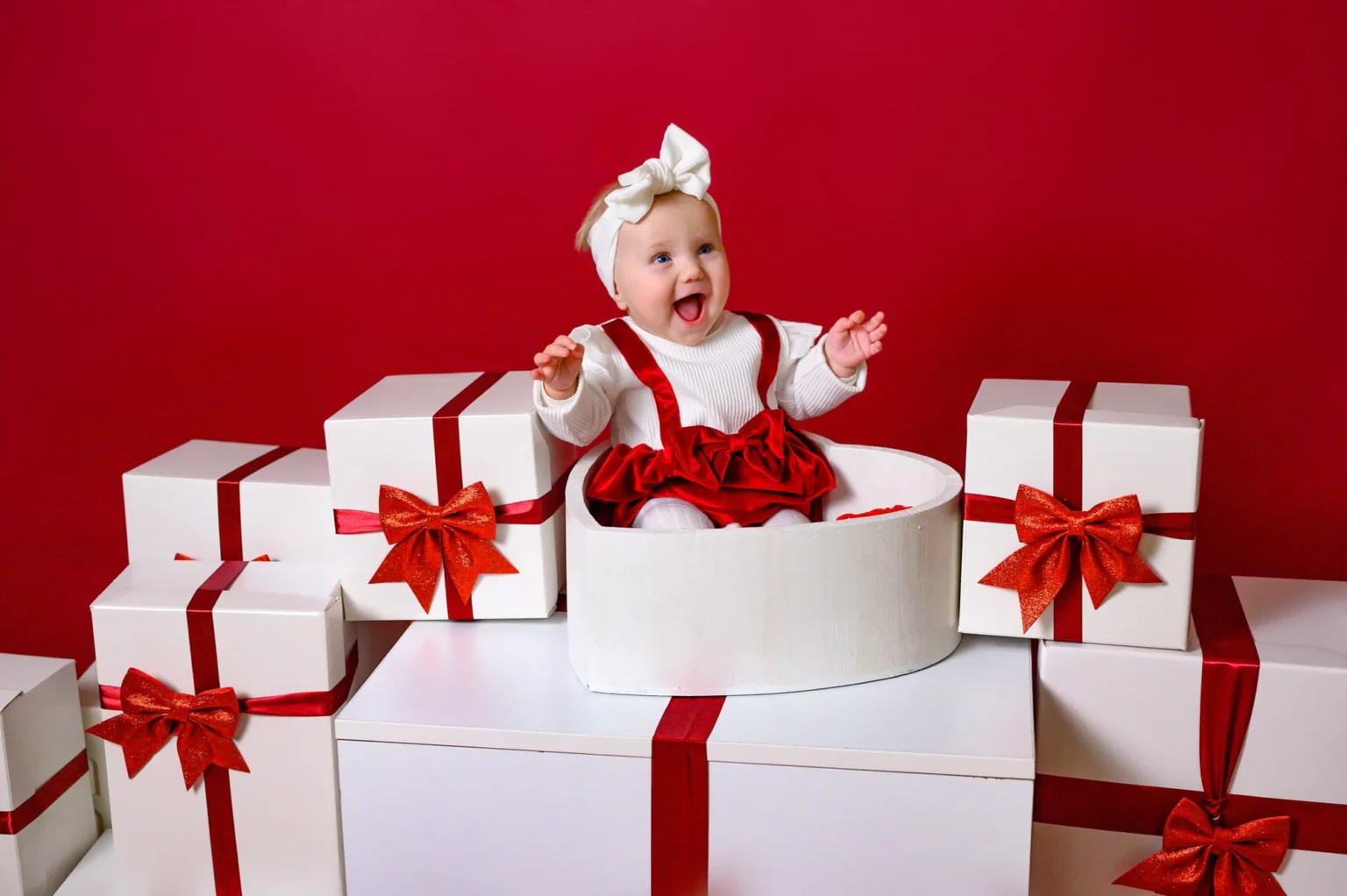 Neonata con fiocco tra pacchi regalo su sfondo rosso, foto di Natale in studio a Venaria Reale